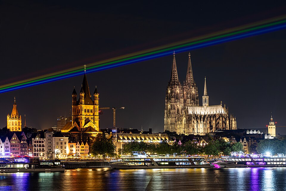 Ein leuchtender Regenbogen überspannt nachts den Rhein vor dem Kölner Dom.
